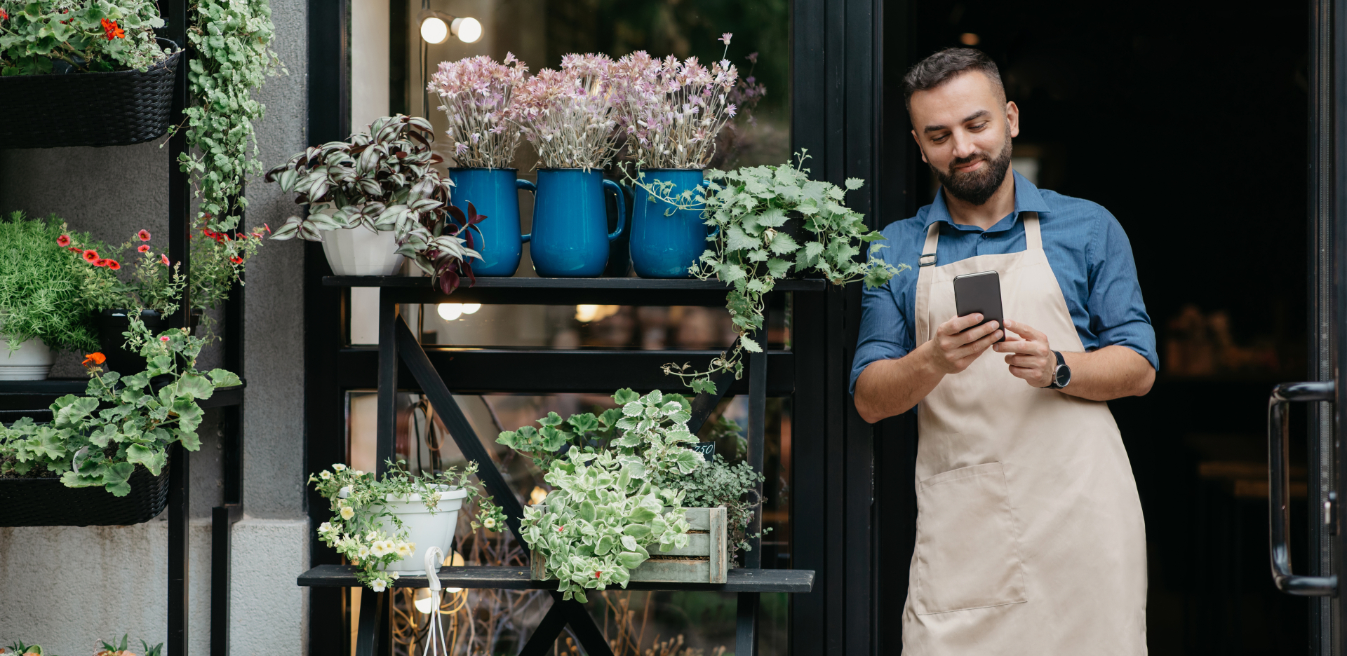 Man leaning against a shelf with potted plants lining the shelves. He is on his phone looking at mobile banking.